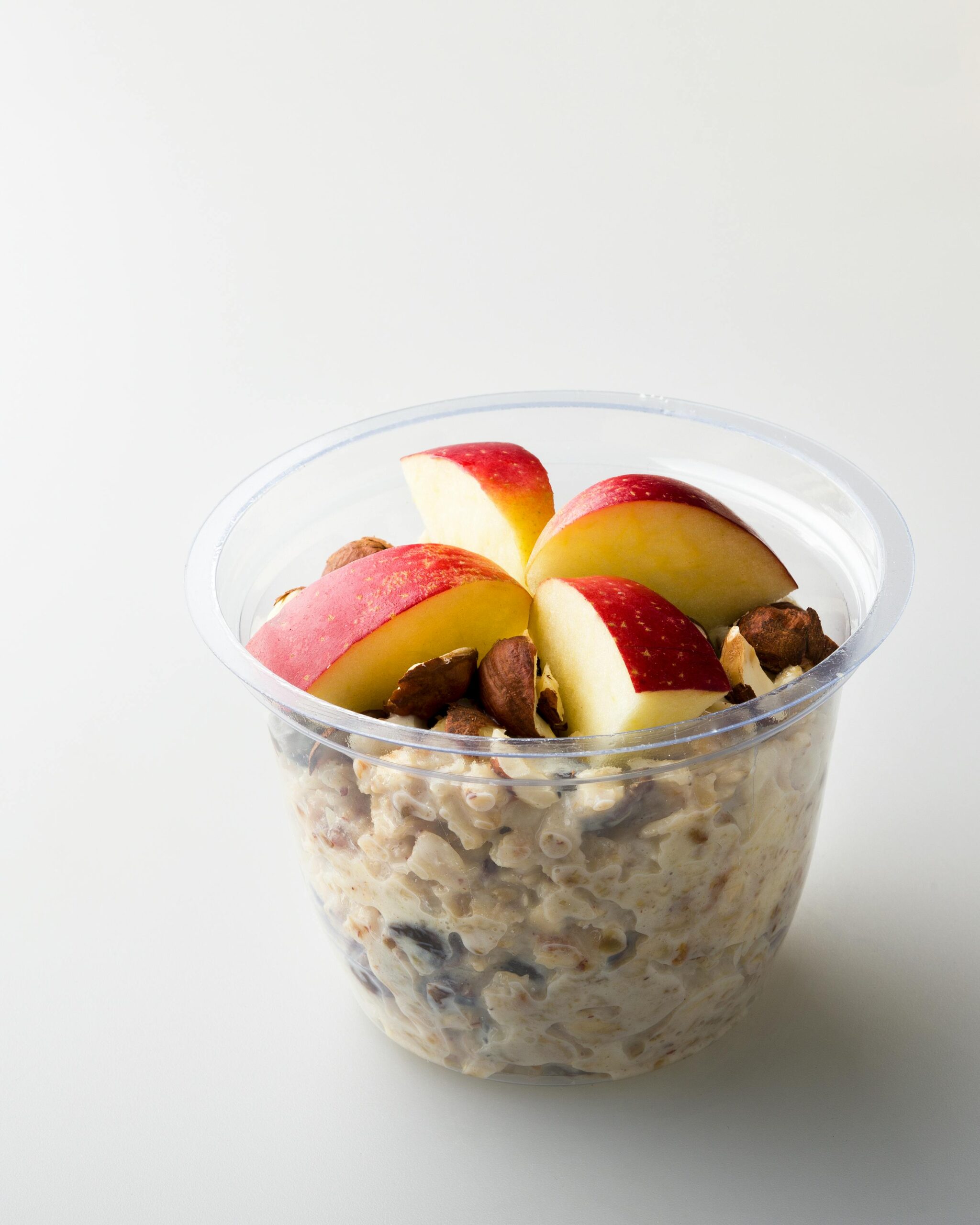 A clear plastic container of oatmeal topped with fresh apple slices and nuts on a white background.